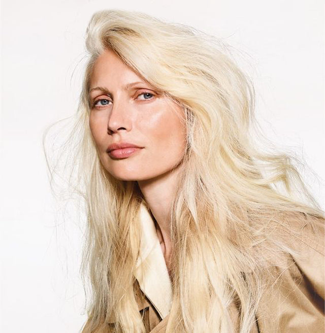 Blonde woman with long, voluminous hair wearing a beige trench coat against a plain background