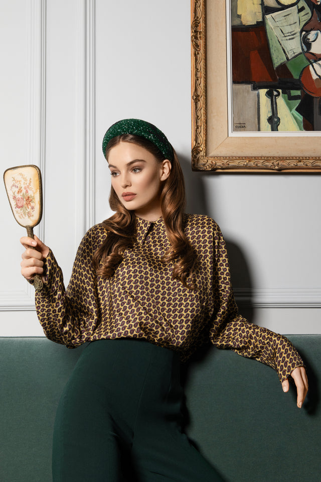 Woman with curled brown hair wearing a green sparkly headband and patterned blouse holding a vintage floral hand mirror