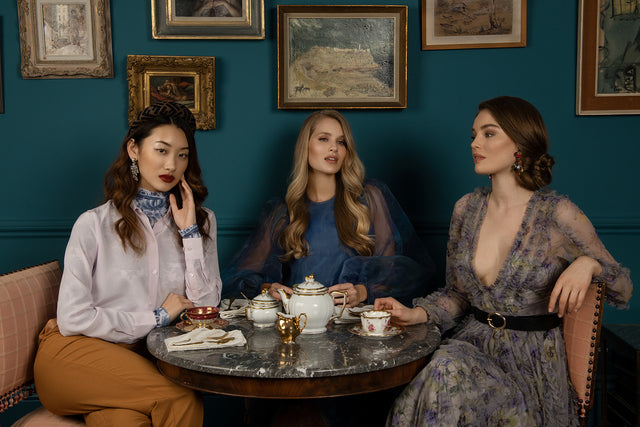 Three women in elegant vintage-inspired dresses seated around a marble table with tea set in a room with teal walls and framed paintings