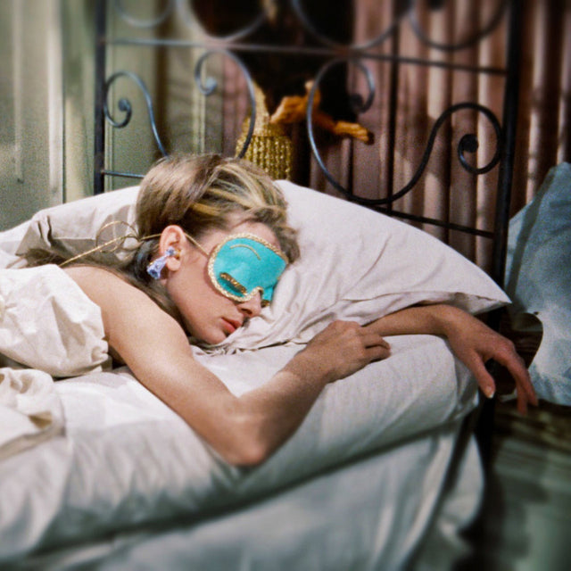 Person lying on bed wearing turquoise sleep mask with gold trim and earplugs, resting on white pillow
