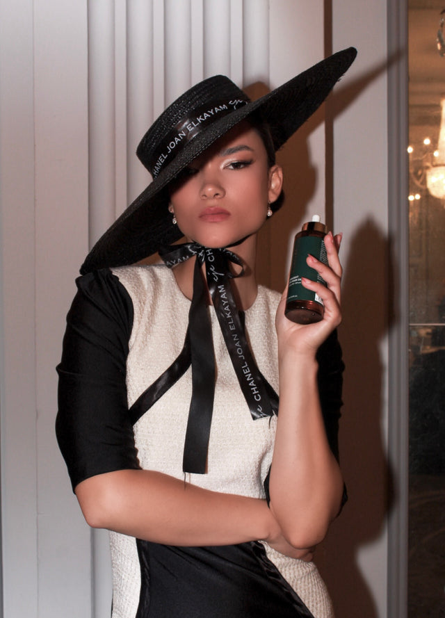 Woman wearing black wide-brimmed hat and black-and-white dress holding green skincare bottle