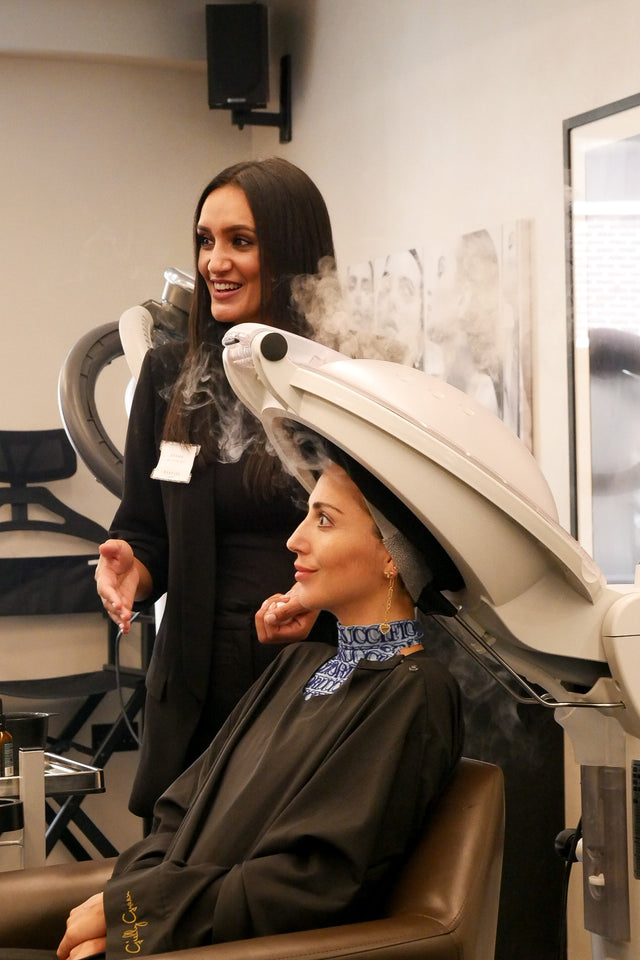 Woman sitting under white hair steamer in salon with stylist standing beside her, both smiling