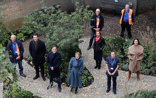 Group of people standing outdoors in autumn coats and uniforms in a garden with gravel paths and greenery