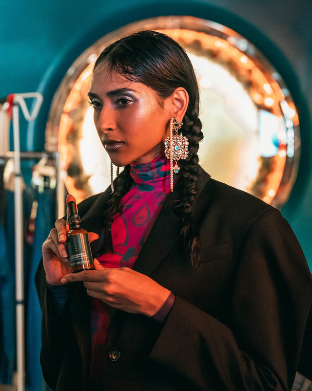 Woman with braided hair wearing statement earrings and black blazer holding amber glass dropper bottle