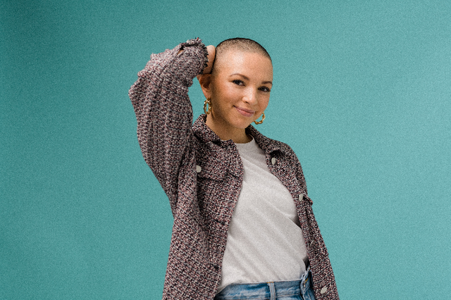 Smiling woman with shaved head wearing gold hoop earrings, white t-shirt, and patterned jacket