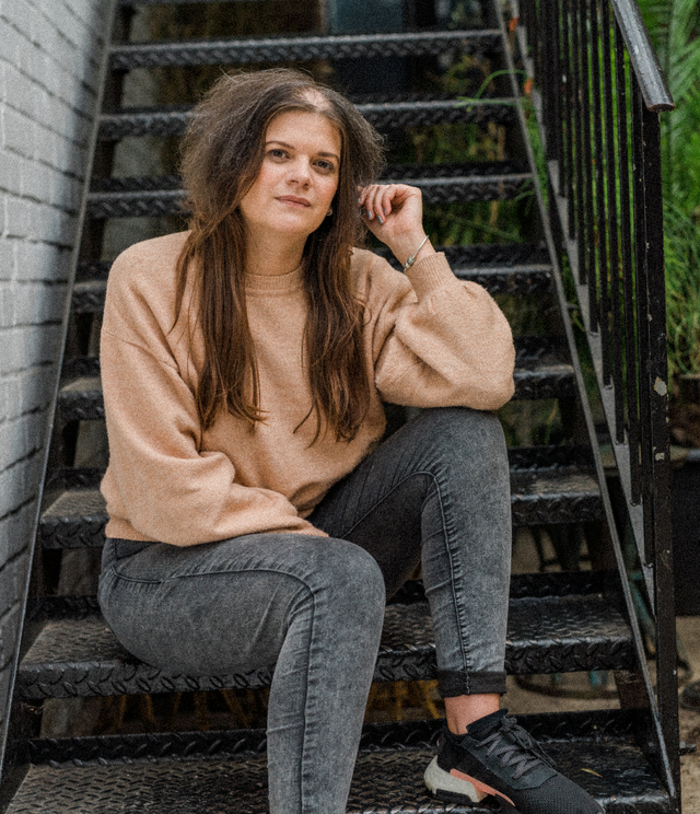 Woman with long brown hair wearing a beige sweater and grey jeans sitting on black metal stairs outdoors