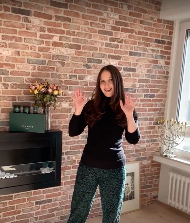Woman with long dark hair wearing black top and patterned green trousers standing indoors