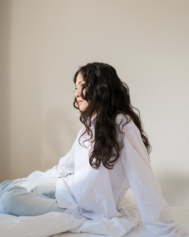 Woman with long wavy dark hair wearing a loose white shirt and light blue jeans sitting on a white bed