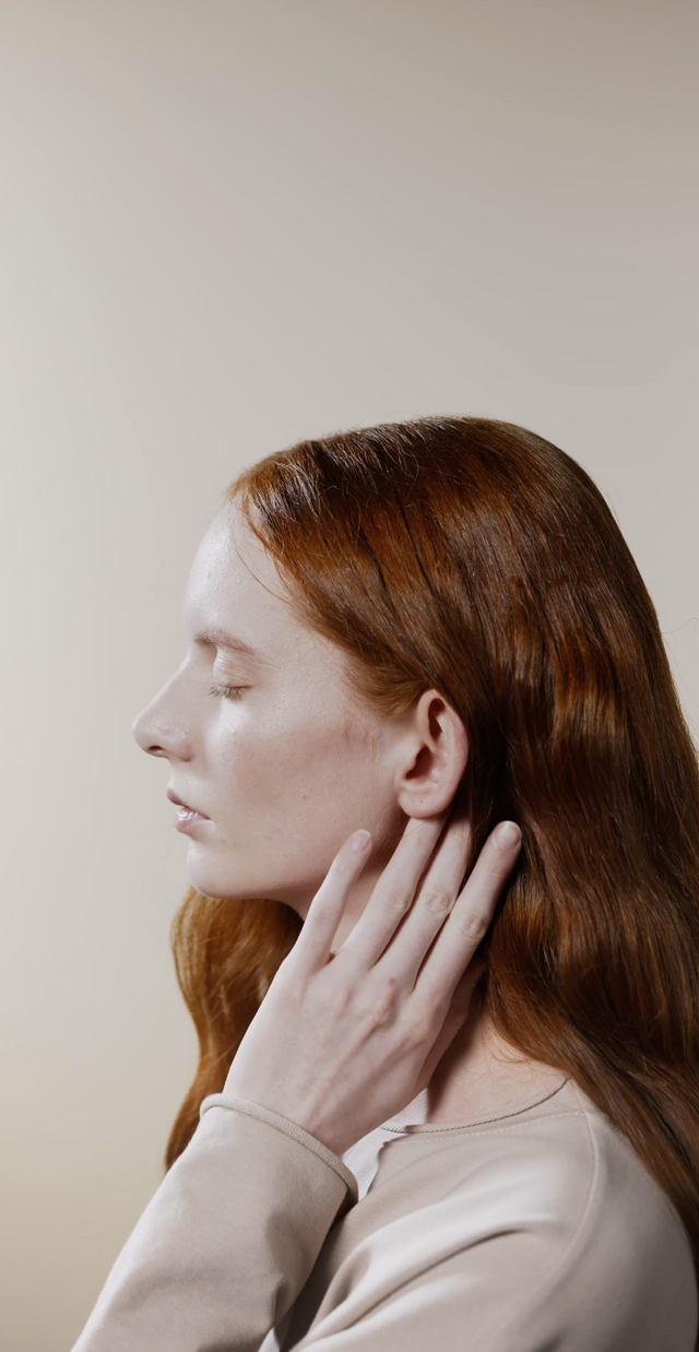 Side profile of a woman with long red hair touching her scalp against a neutral background