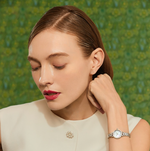Woman with sleek brown hair and red lipstick wearing a white top and silver wristwatch against green background