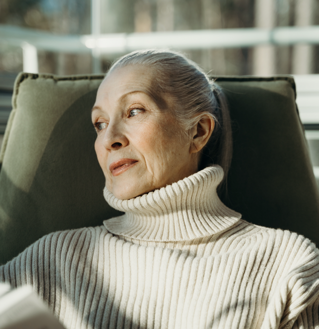 Older woman with silver hair in a cream ribbed turtleneck sweater sitting on a green chair