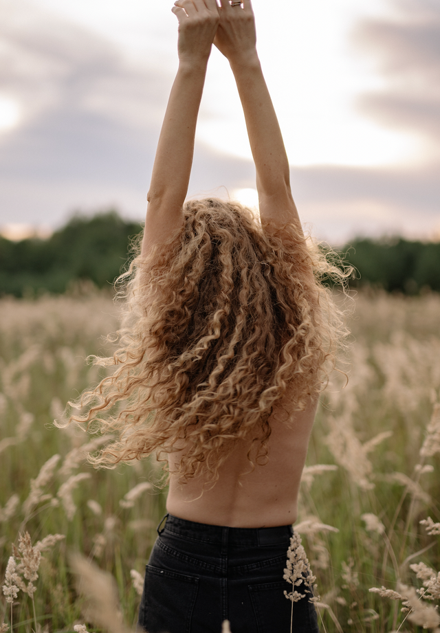 Person with long, curly blonde hair standing in a grassy field at sunset with arms raised