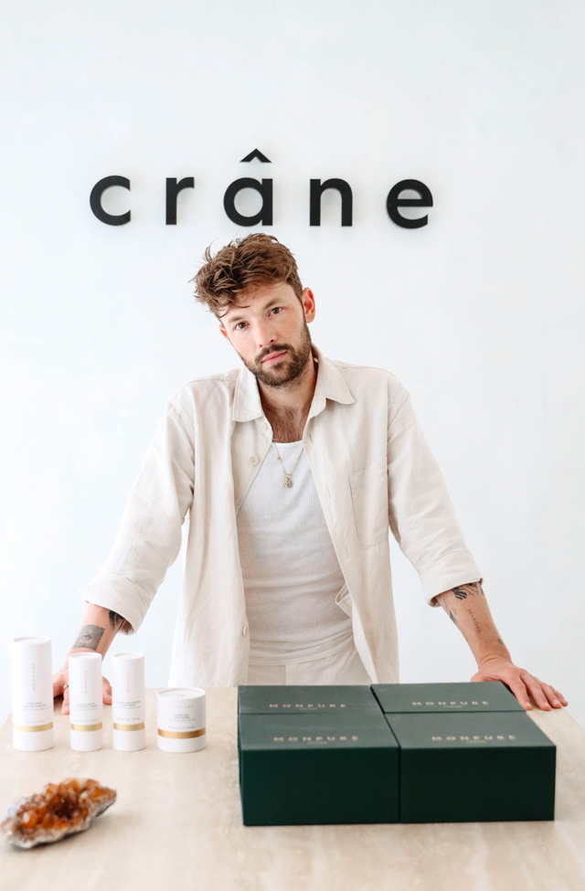 Man with tousled hair wearing cream shirt standing behind table with skincare products and dark green boxes