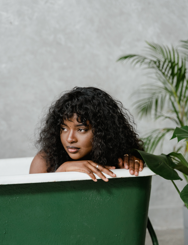 Woman with long curly black hair resting on edge of green bathtub with indoor plants nearby