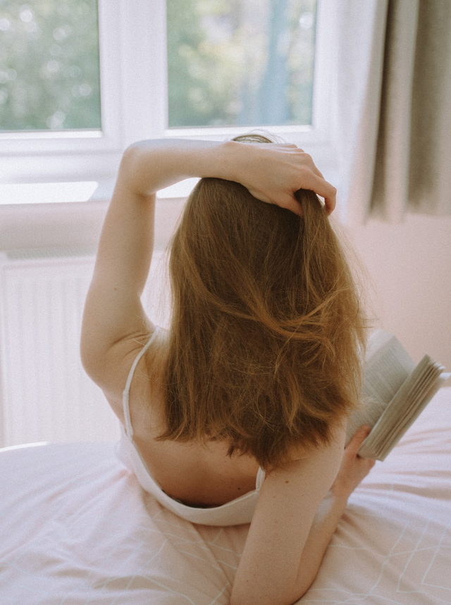 Person with medium-length brown hair lying on bed reading a book near a window with natural light