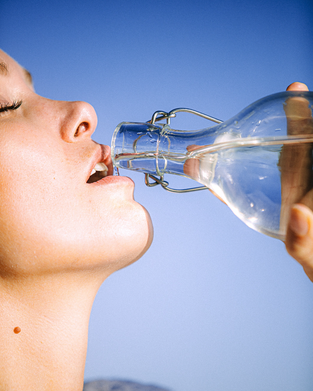 Close-up of person drinking clear water from a glass bottle against a blue sky