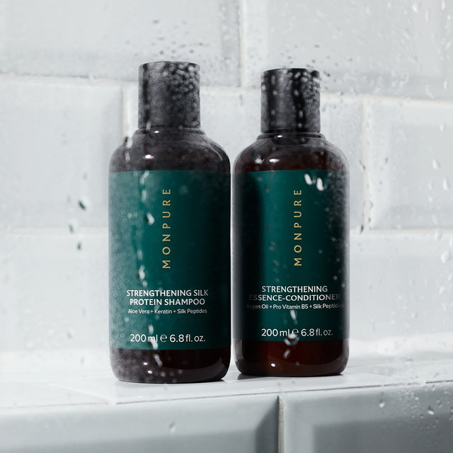 Two brown bottles of MONPURE strengthening hair care products with dark green labels on white tiled surface with water droplets