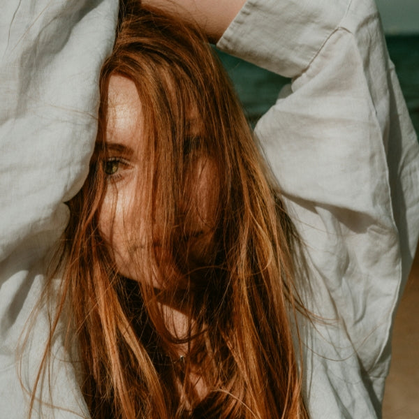 Woman with long auburn hair wearing a loose white linen shirt, arms raised above head