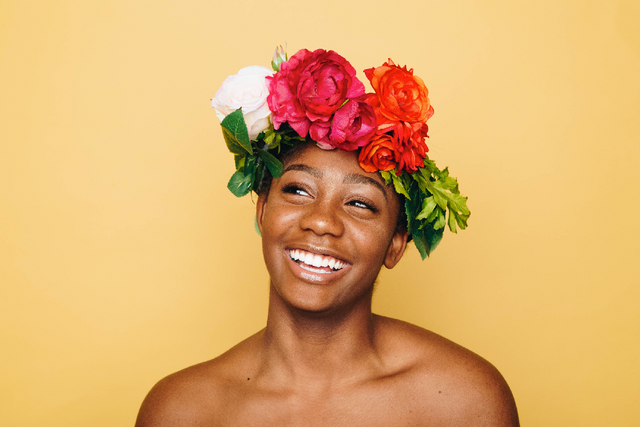 Smiling woman wearing a vibrant floral crown with red, pink, and white flowers against yellow background