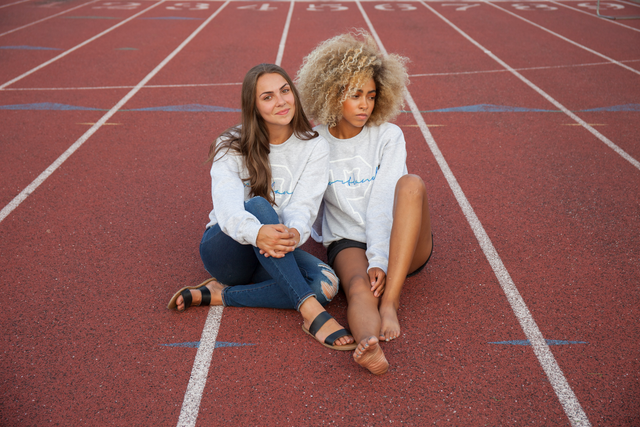 Two young women sitting on a red running track wearing matching light grey sweatshirts, one with curly blonde hair and the other with long straight brown hair