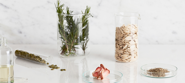 Clear glass containers with green herbs, seeds, pink mushroom, and ground spices on white surface