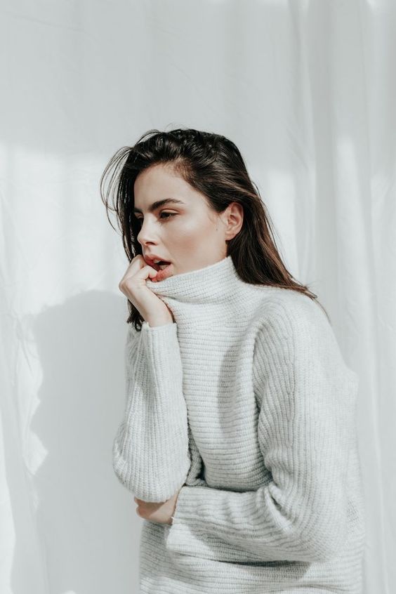 Woman with long dark hair wearing a light grey ribbed turtleneck sweater against a white background