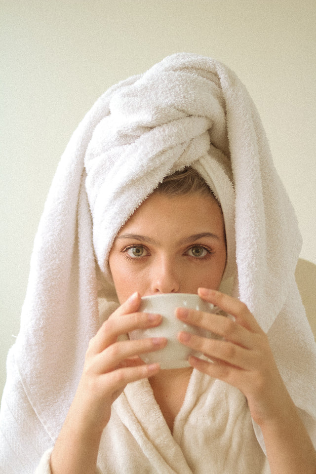 Woman with white towel wrapped around hair and wearing bathrobe, holding white cup near face