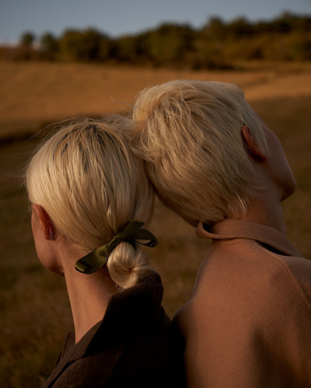 Two people with fine blonde hair styled simply, one with a green ribbon, outdoors in warm light
