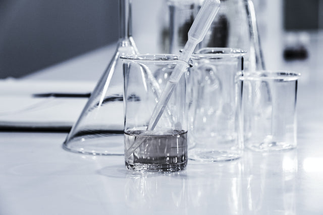 Clear glass beakers and a pipette with liquid on a reflective white surface in a laboratory setting
