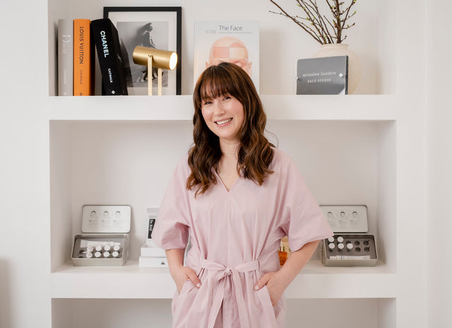 Smiling woman in a light pink belted robe standing in front of white shelves with skincare products and books