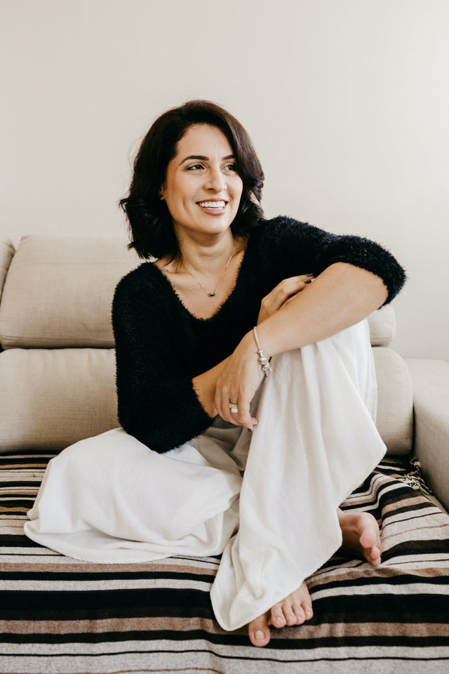 Smiling woman with dark hair wearing a black fuzzy sweater and white wide-leg trousers sitting barefoot on striped fabric