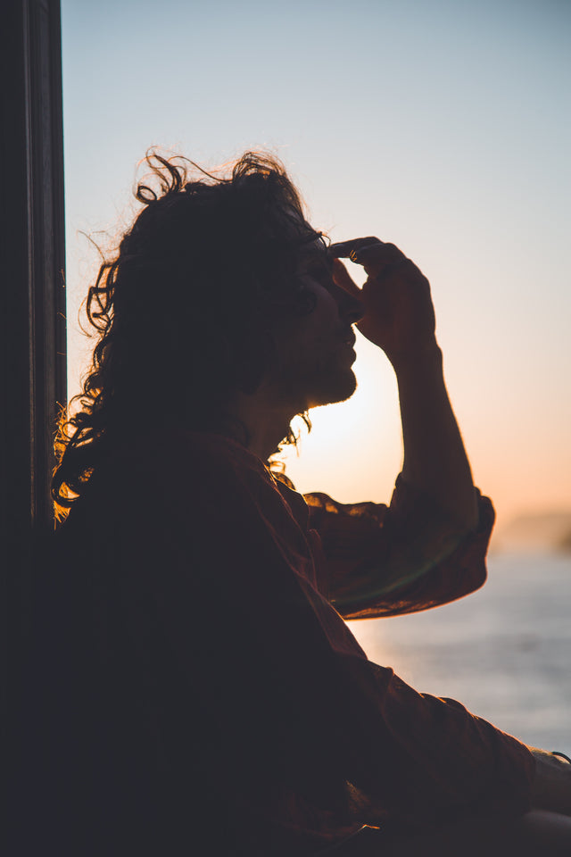 Silhouette of person with curly hair touching forehead, sitting by window at sunset