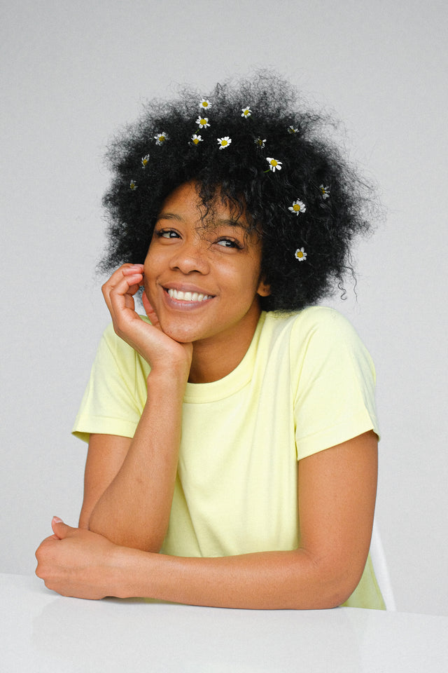 Smiling woman with natural curly hair adorned with small white daisies, wearing a pale yellow t-shirt