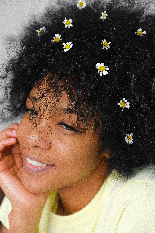Smiling woman with natural curly hair adorned with small white and yellow daisies, wearing a pale yellow top