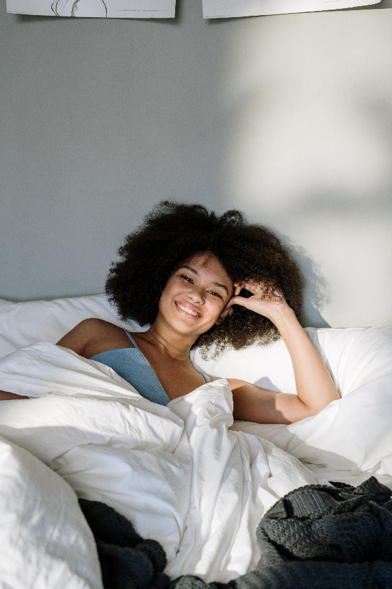 Smiling woman with natural curly hair lying in bed under white duvet, wearing blue top