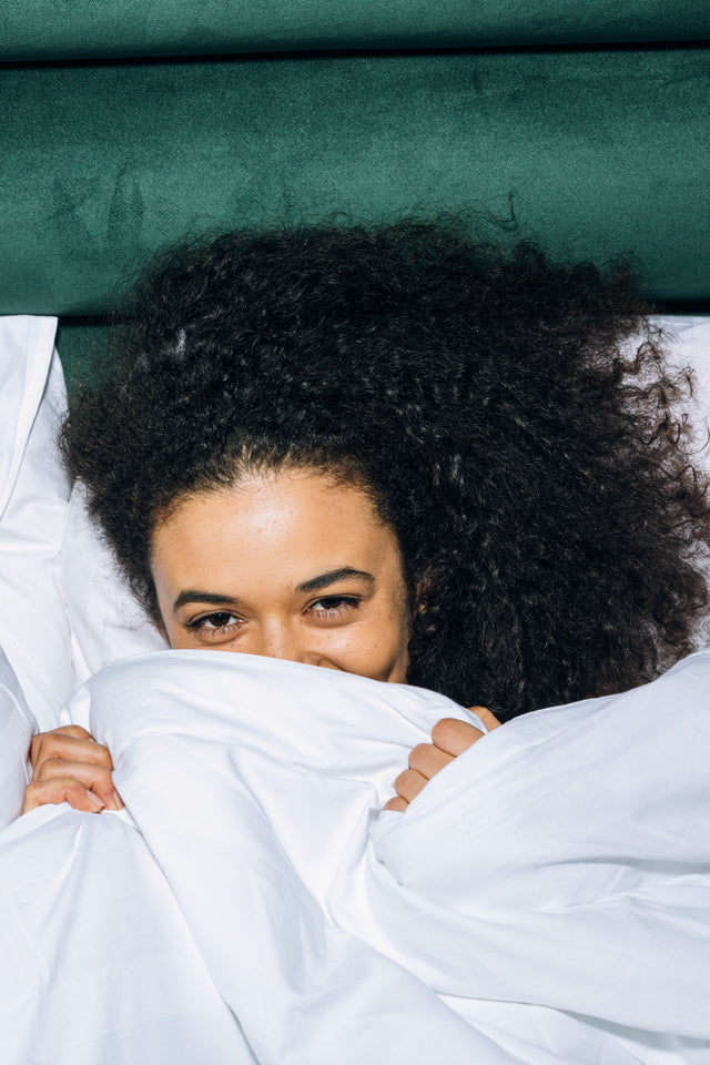 Person with voluminous curly black hair peeking from behind white bedding on green upholstered headboard