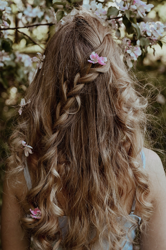 Long wavy light brown hair with a loose braid and pink flowers, outdoors in nature