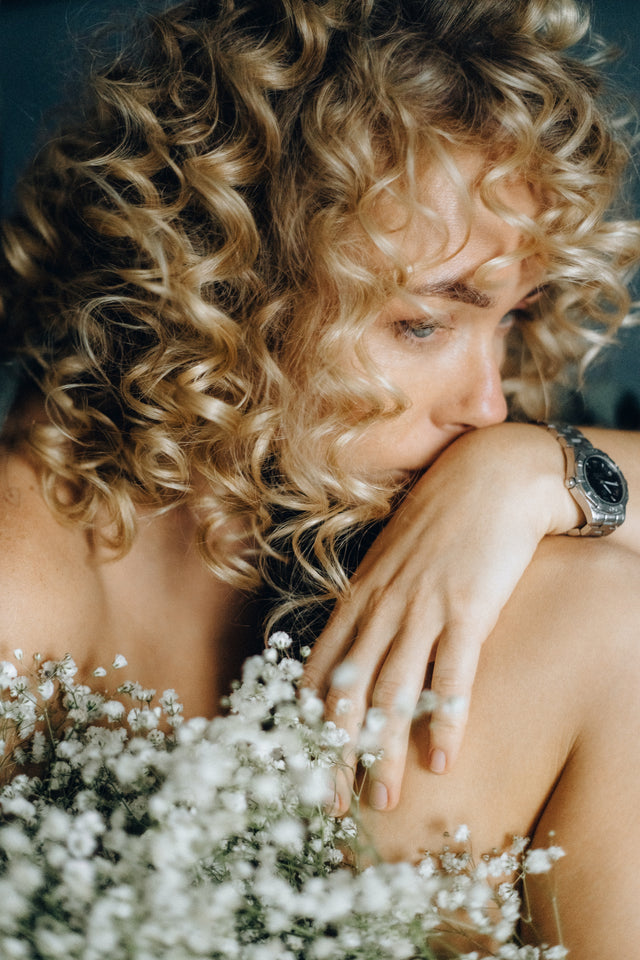 Blonde woman with curly hair resting chin on knee, surrounded by delicate white flowers
