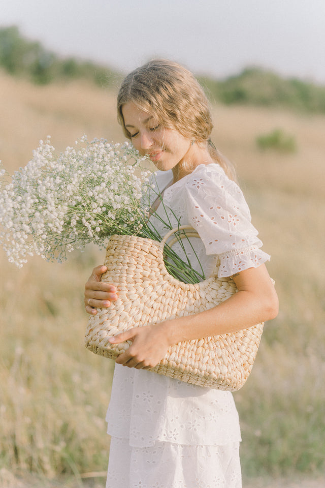 Woman in white eyelet dress holding woven straw handbag with white baby's breath flowers