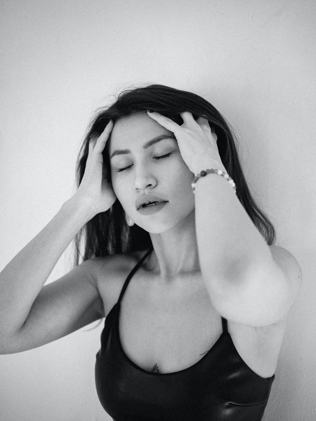 Woman with long dark hair wearing a black sleeveless top, touching her scalp with closed eyes