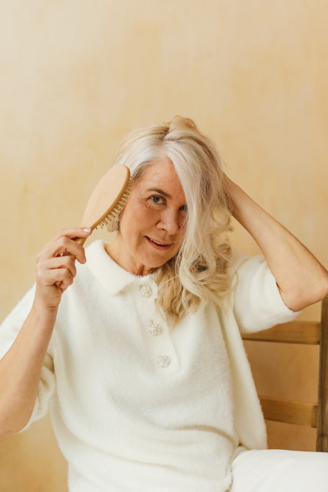 Mature woman with long silver hair brushing hair with wooden hairbrush wearing white sweater