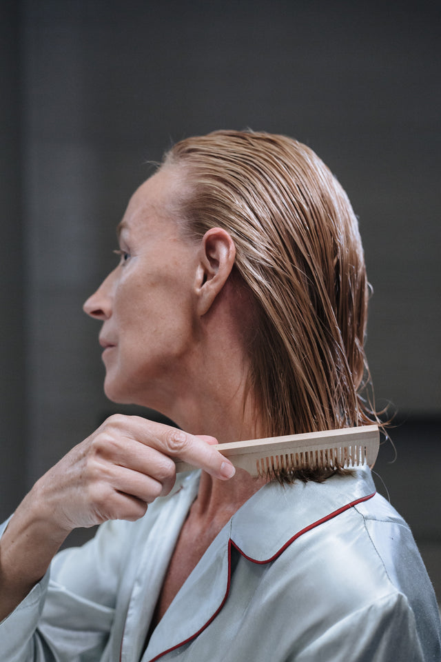 Woman with wet blonde hair combing with wooden comb wearing light blue satin pyjamas