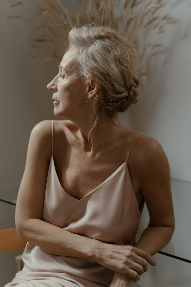 Mature woman with grey hair styled in a loose bun wearing a pale pink satin camisole