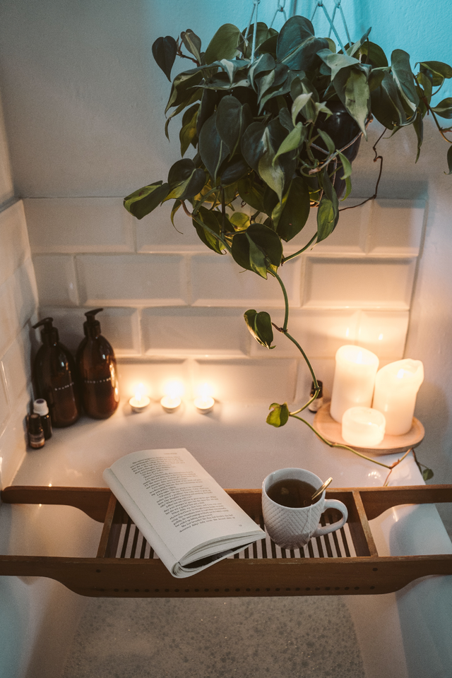 Wooden bath tray holding an open book and a white cup of tea, surrounded by candles and dark glass bottles
