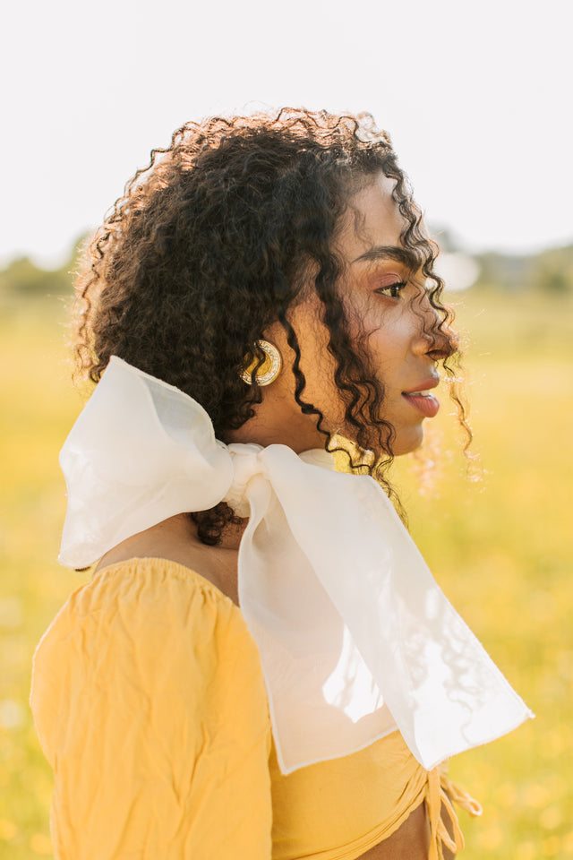 Woman with curly hair wearing large white bow scarf and yellow off-shoulder top outdoors