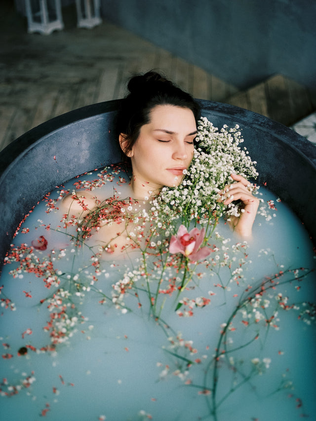 Woman relaxing in a milky bath with white and pink flowers, holding a bouquet close