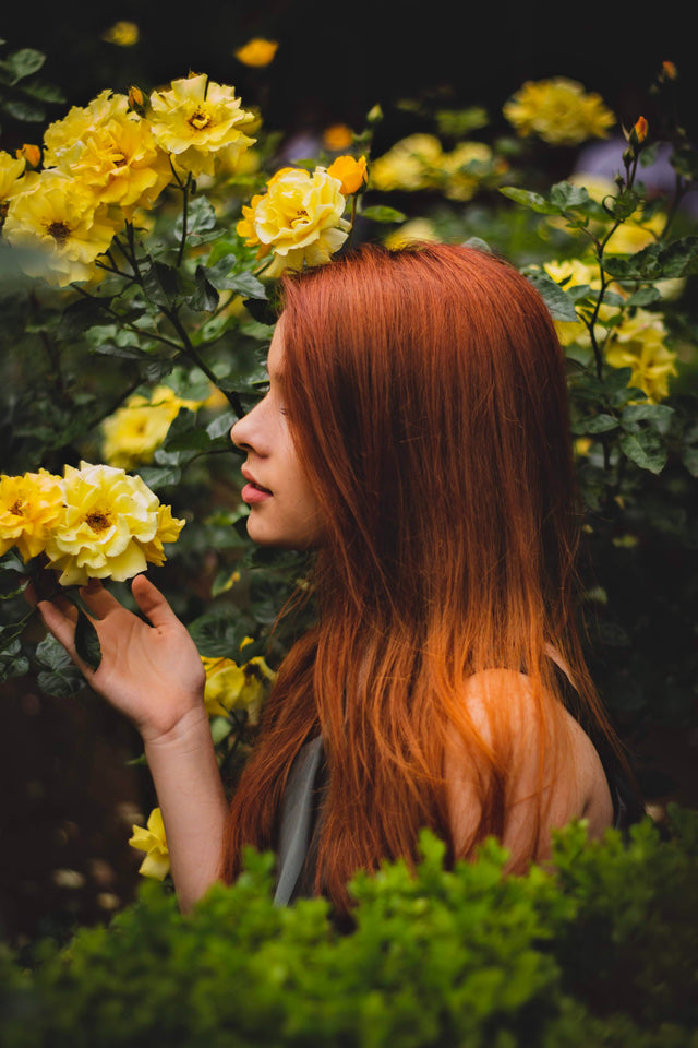 Woman with long vibrant red hair gently touching yellow roses in a lush garden