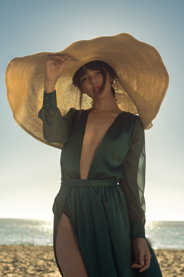 Woman wearing large wide-brimmed straw sun hat and deep green sheer long-sleeve dress on beach