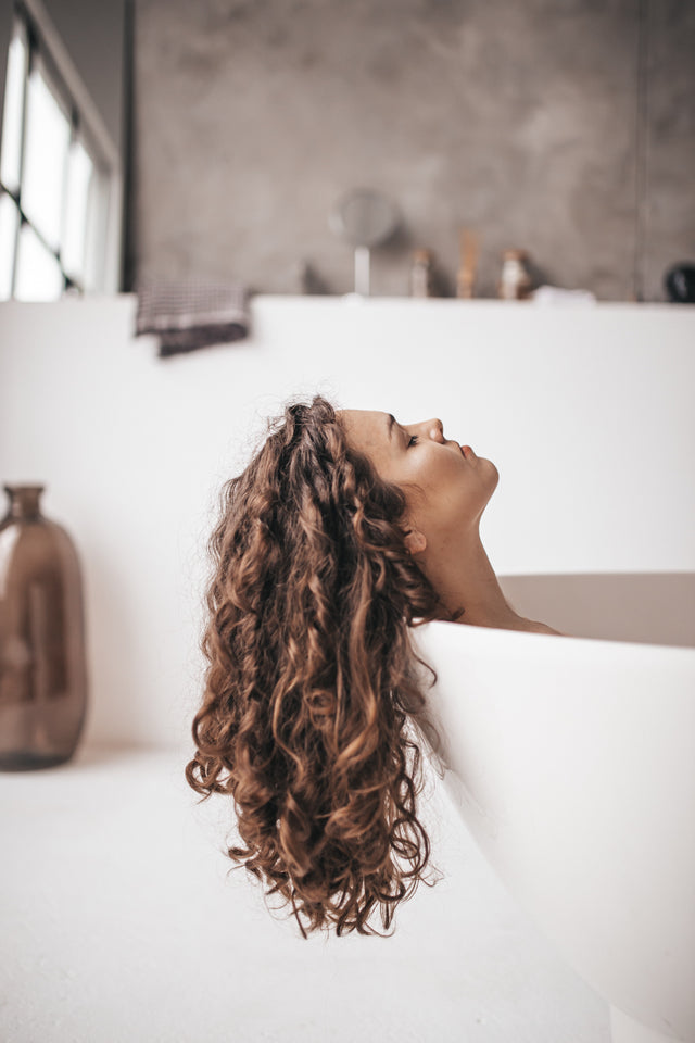 Woman with long curly brown hair relaxing in a modern white bathtub in a minimalist bathroom