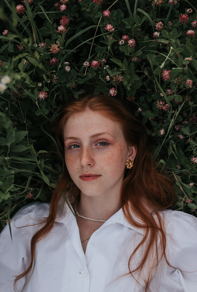 Young woman with long red hair lying in green grass with small pink flowers, wearing white shirt and gold earrings